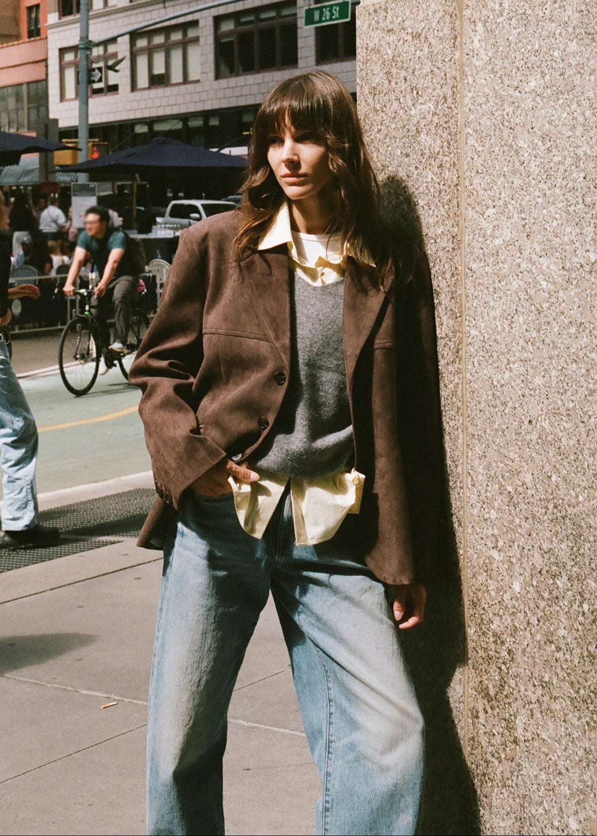 A woman wearing a brown brown suede jacket over a gray sweater and butter color cotton button down shirt.