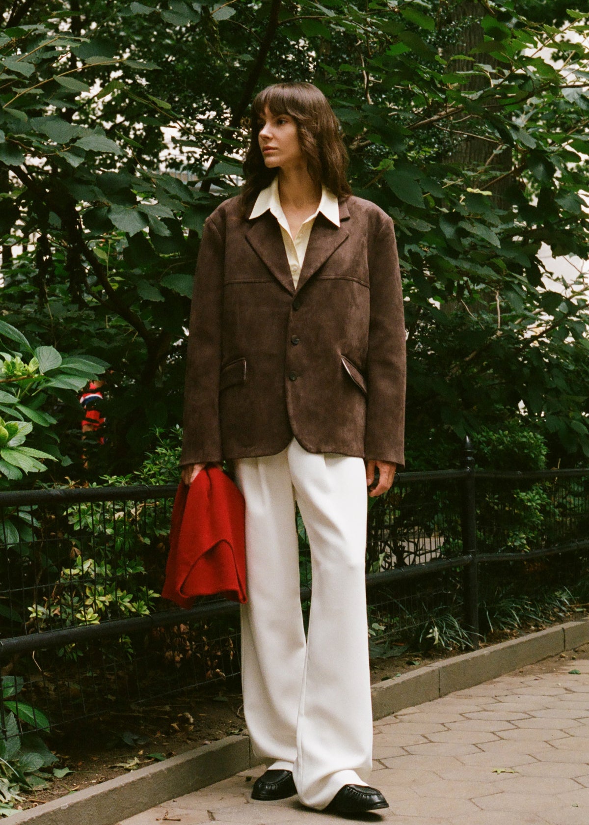 A woman stands outdoors on a paved walkway in front of dense green trees, wearing an oversized brown jacket over a cream shirt, white wide-leg pants, and black shoes, while holding a bright red sweater.