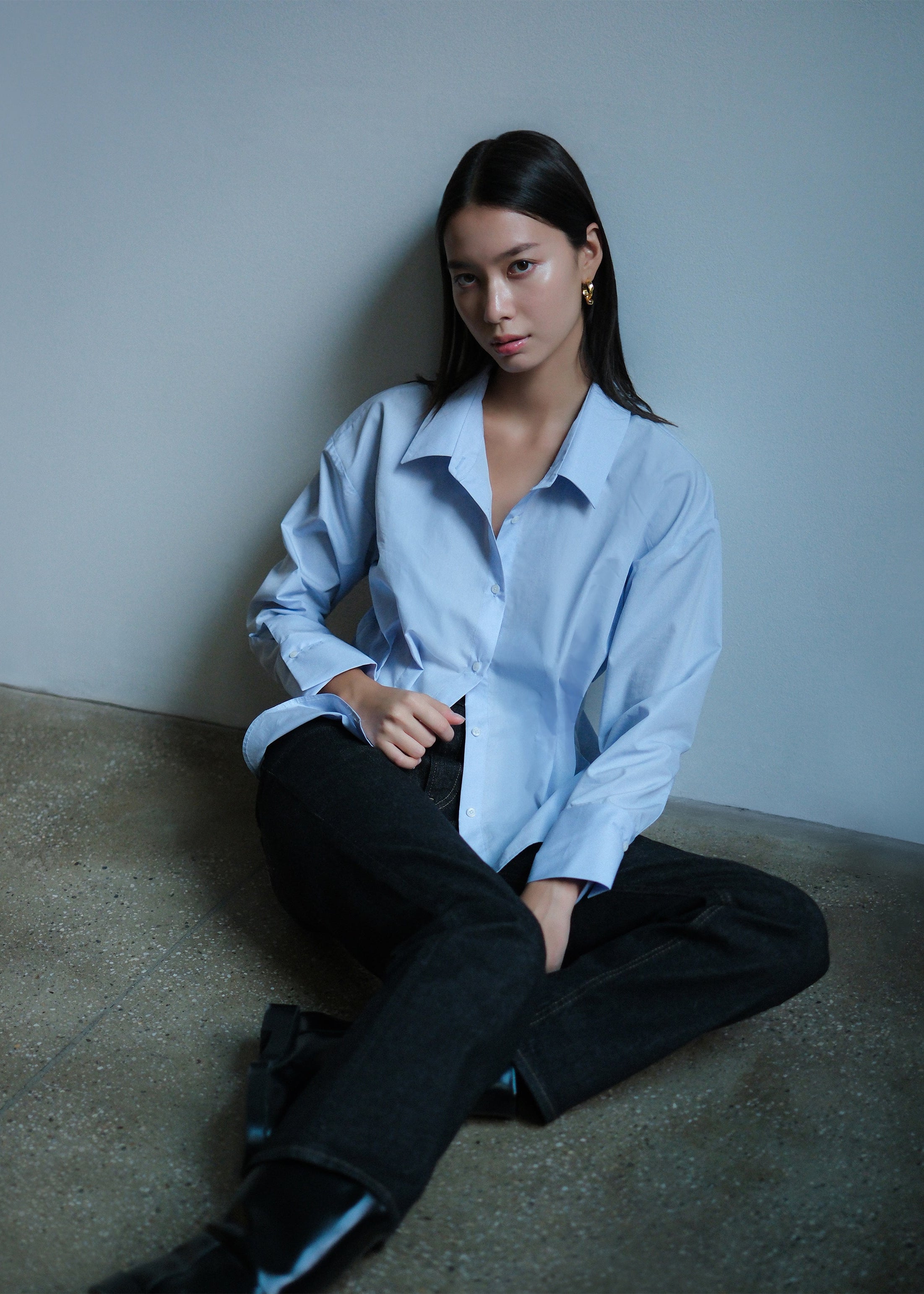 Woman wearing a light blue shirt and dark pants sitting on a concrete floor against a plain wall.