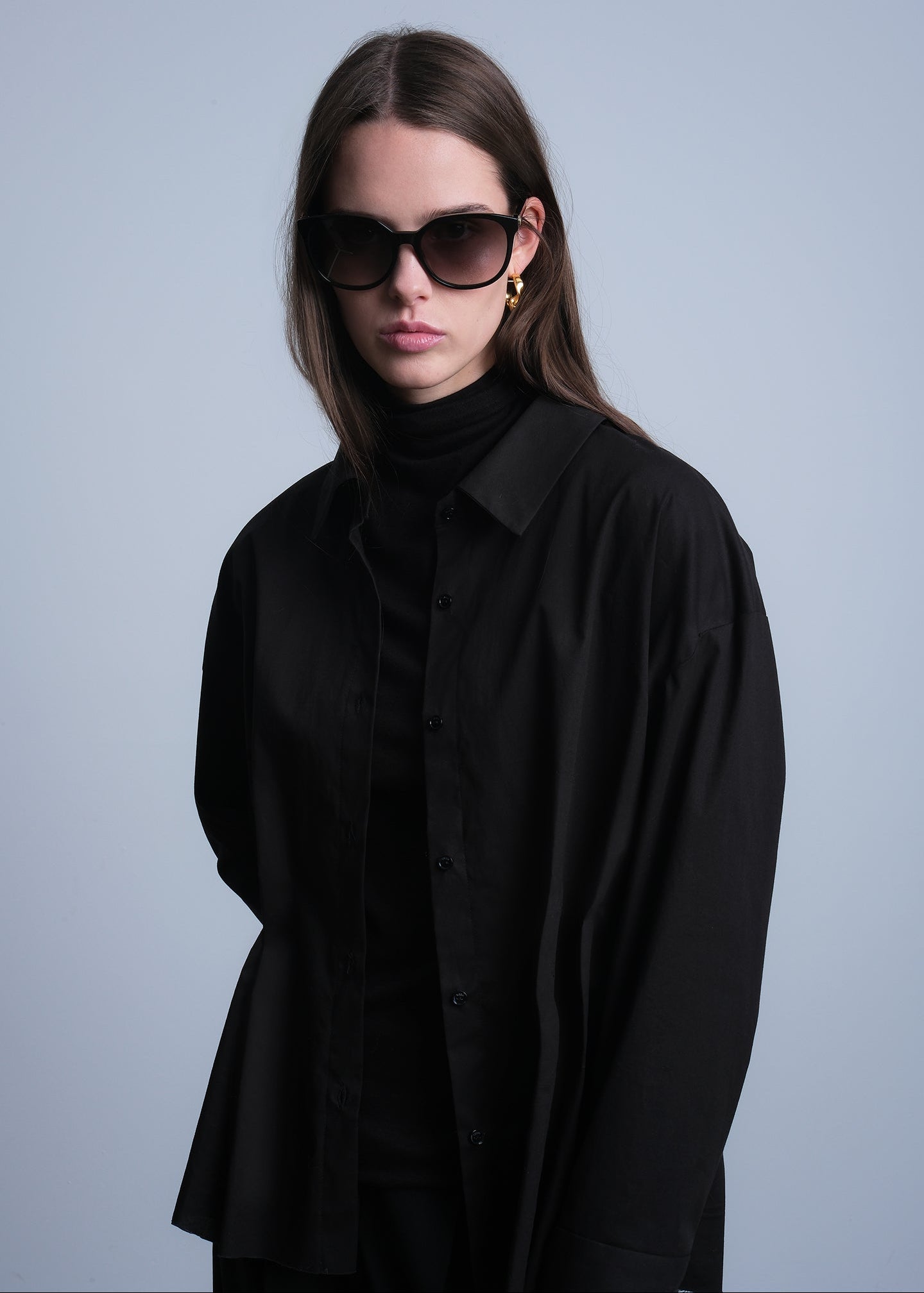 Woman wearing a black shirt with pleated detail on the waist and sunglasses against a gray background