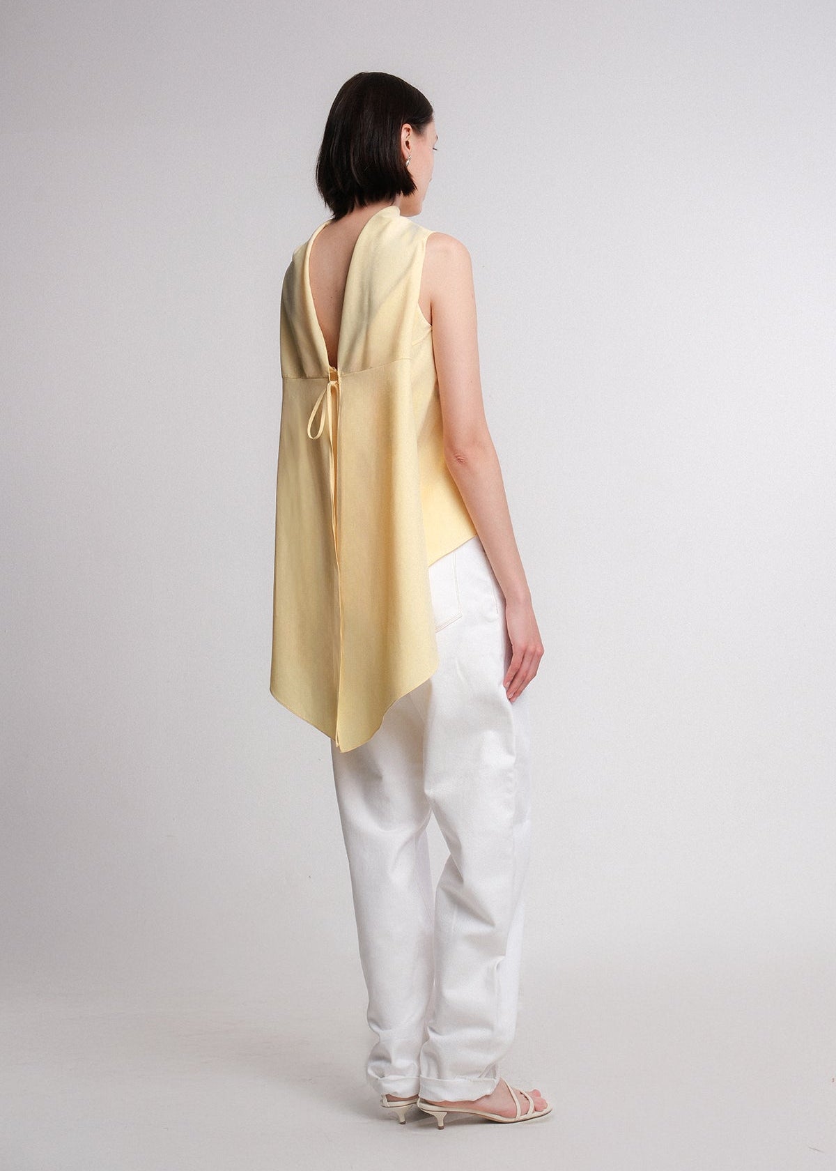 Sleeveless butter yellow cotton-rayon-poly blend top with an asymmetrical high-low hem and extended draped back panel tied at the side, featuring a clean crew neckline and soft, fluid silhouette.