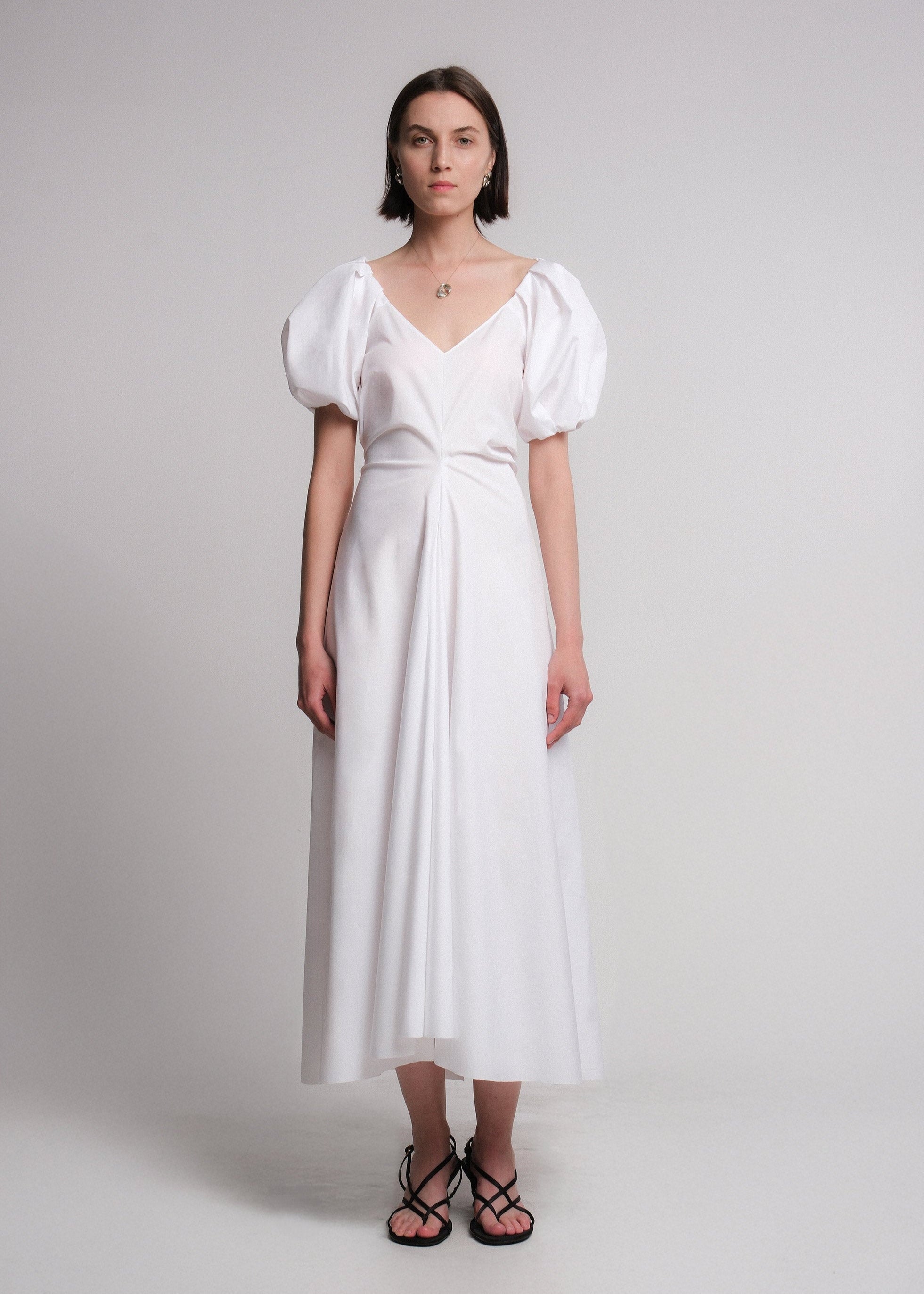 Maxi dress made of white cotton poplin fabric, featuring voluminous puff sleeves, a deep V-neckline, a gusset for added movement, and a flared A-line skirt.