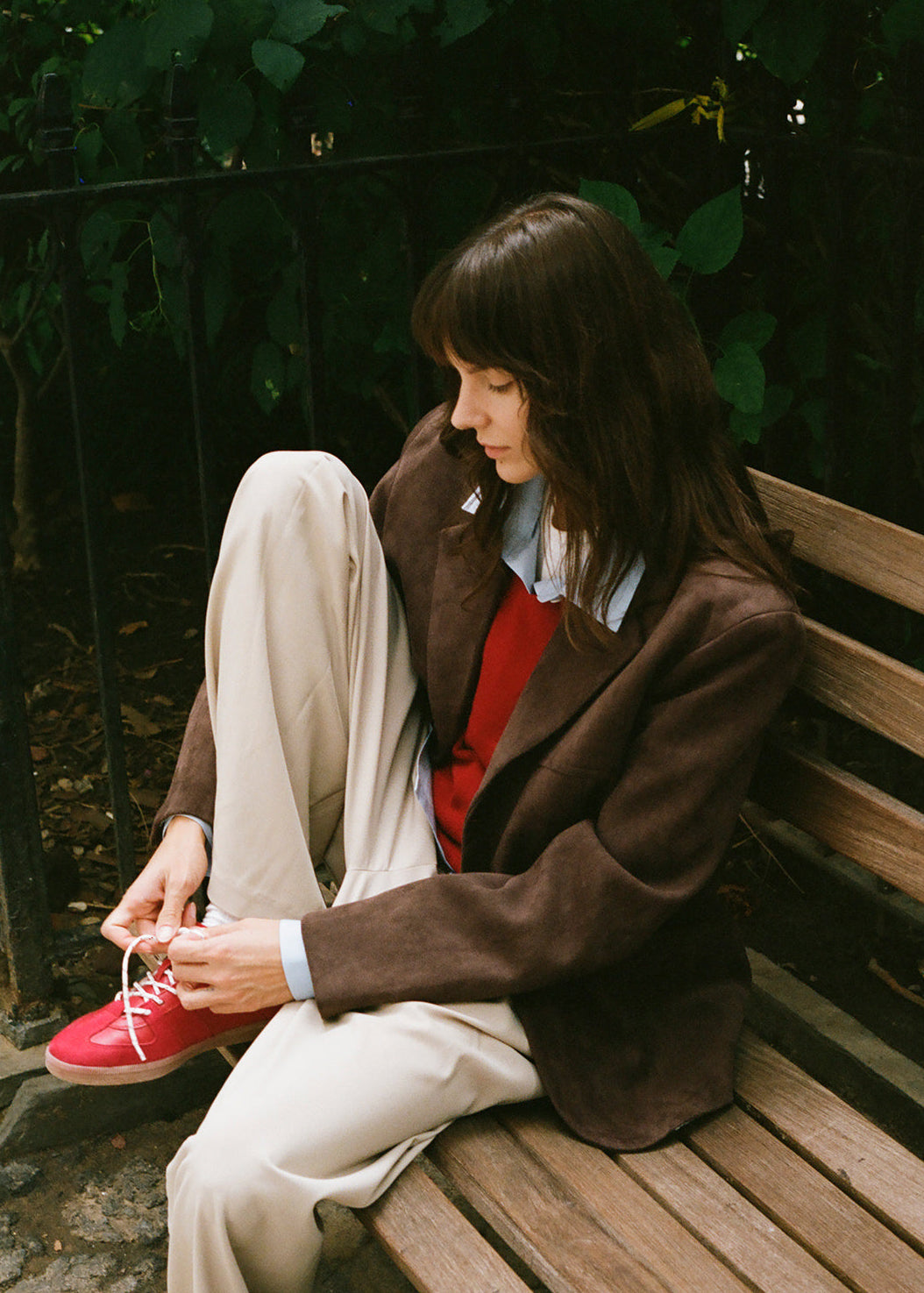 She is wearing a brown faux suede blazer, cream trousers, a red top, and a light blue collared shirt.