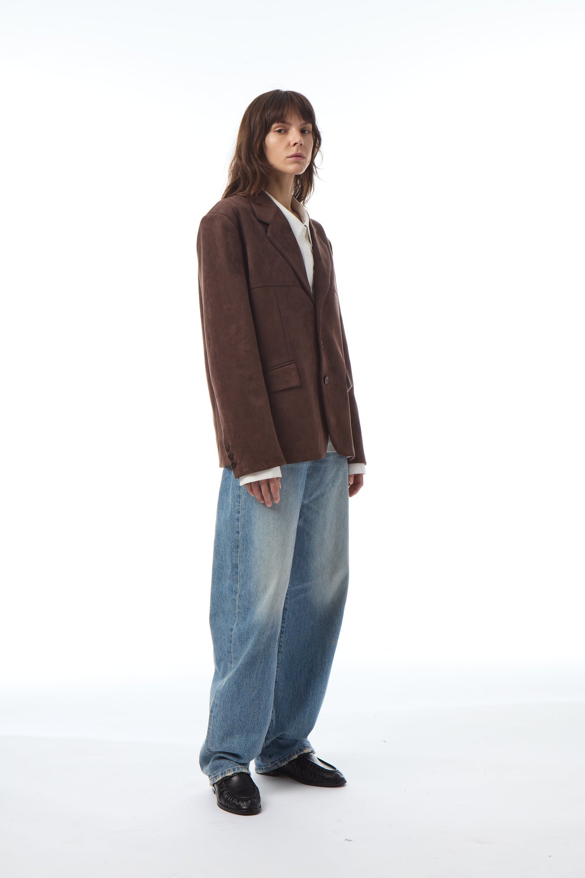 A model stands in a studio wearing a brown corduroy blazer over a white shirt, paired with wide-leg light blue jeans and black shoes, against a solid white background.