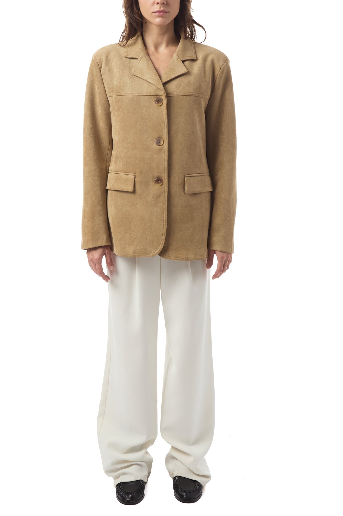 A model stands against a white background wearing a tan suede blazer with flap pockets and white wide-leg trousers.