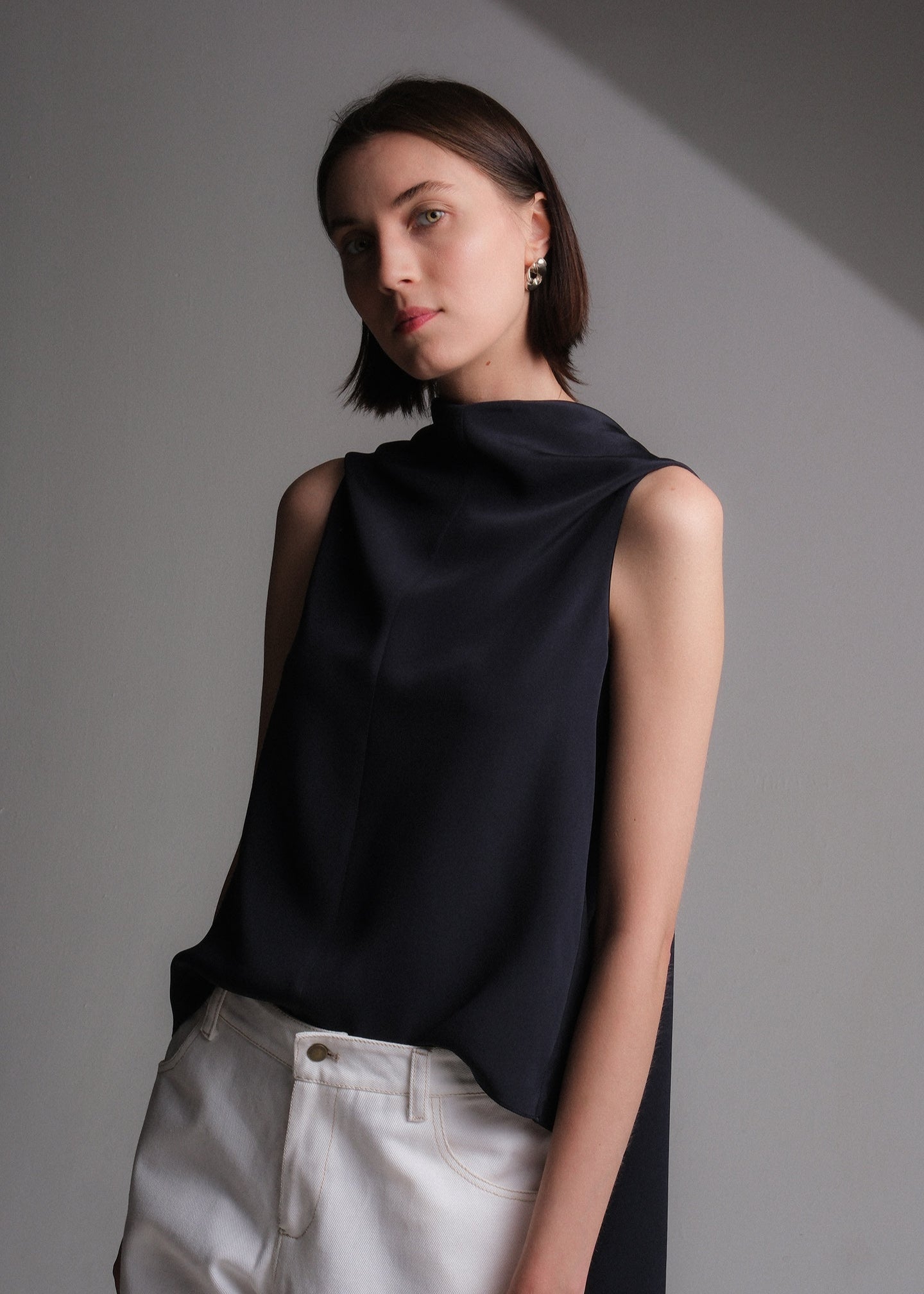 Sleeveless navy silk top with an asymmetrical high-low hem and extended draped back panel tied at the side, featuring a clean crew neckline and soft, fluid silhouette.