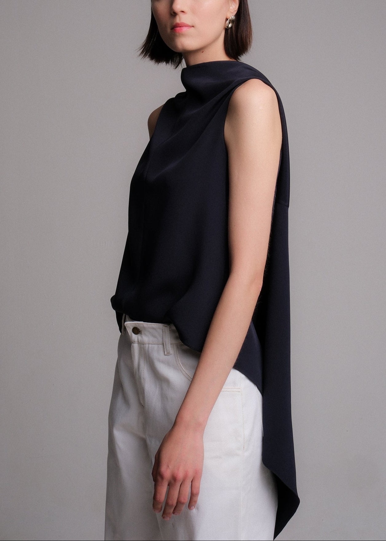 Sleeveless navy silk top with an asymmetrical high-low hem and extended draped back panel tied at the side, featuring a clean crew neckline and soft, fluid silhouette.